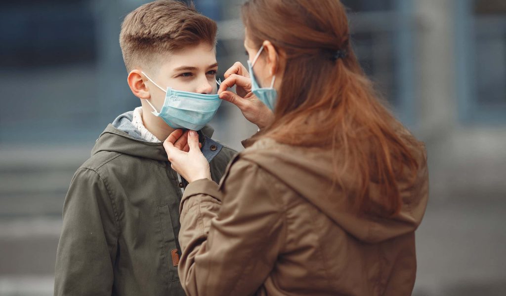 A boy and mother are wearing protective masks. The kid is sneezing and mother is helping him to wear a piece of protective equipment.